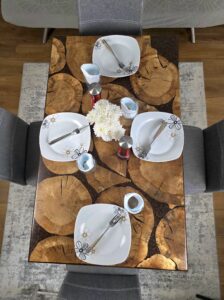 A unique and handcrafted epoxy resin table. The tabletop features a mosaic of irregularly shaped, light brown wood slices embedded within a dark brown, glossy epoxy resin. Some of the wood slices display visible tree rings and natural cracks. The resin is smooth and reflective, catching the light. The table has a dark brown wooden edge and rests on sturdy black legs. The table is a stylish, nature-inspired piece of furniture with a modern touch.